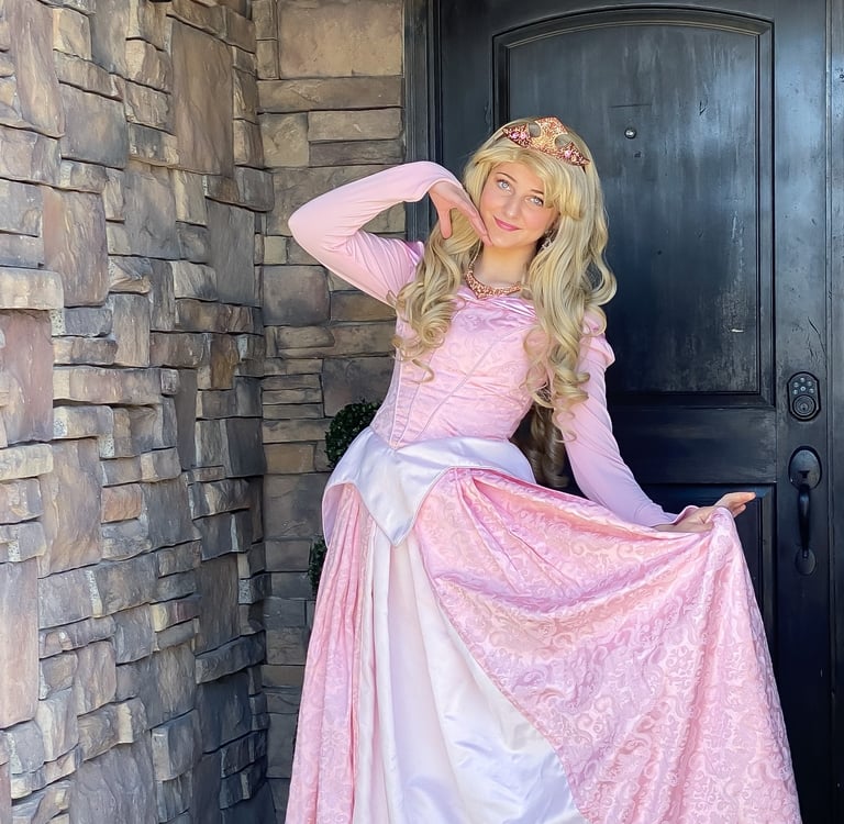 A blonde princess performer in a pink Aurora dress and crown poses by a stone wall for a birthday party.
