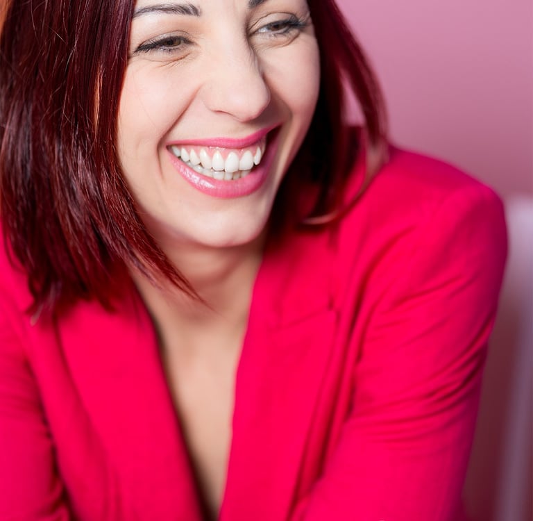 Portrait d’une comédienne qui rit, en veste rose sur fond rose, sourire naturel et expressif.