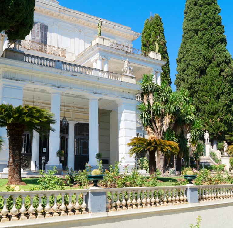 The white Neoclassical facade of Achilleion Palace in Corfu, Greece, surrounded by lush gardens and cypress trees.