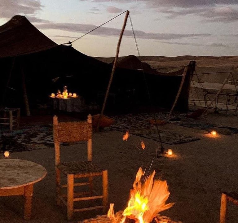 A glowing campfire at a luxury desert camp in Morocco with traditional tents and seating at dusk.