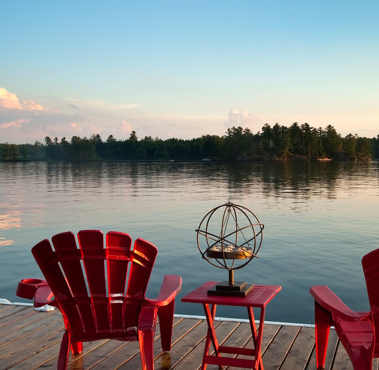 Red Adirondack chairs on a wooden lake dock at sunset with a scenic forest background.