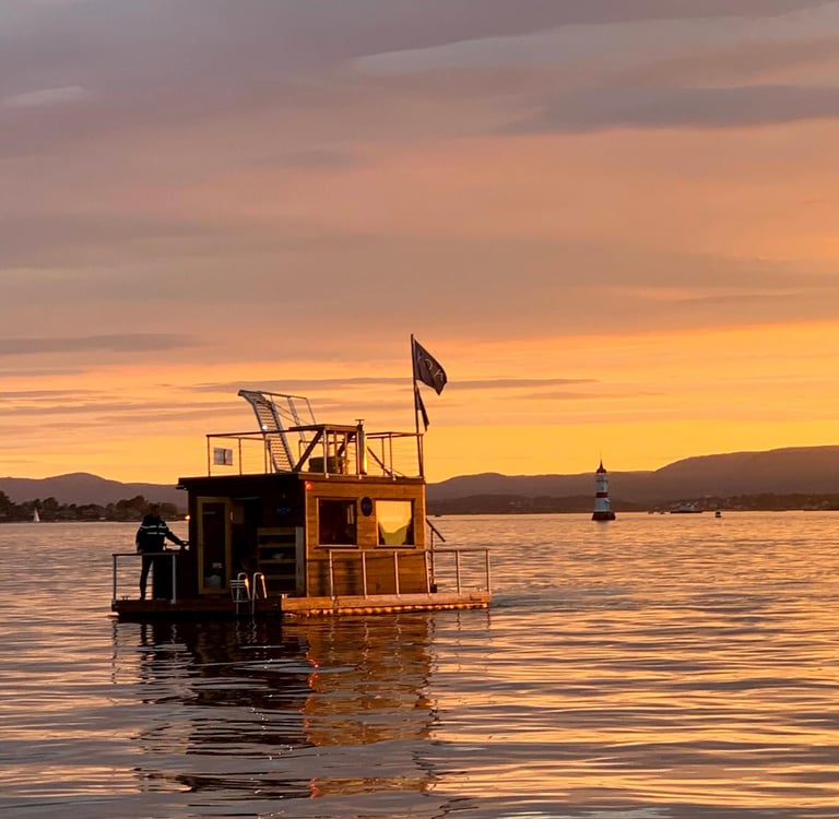 A small wooden houseboat floats on calm water during a golden sunset near a distant lighthouse and hills.