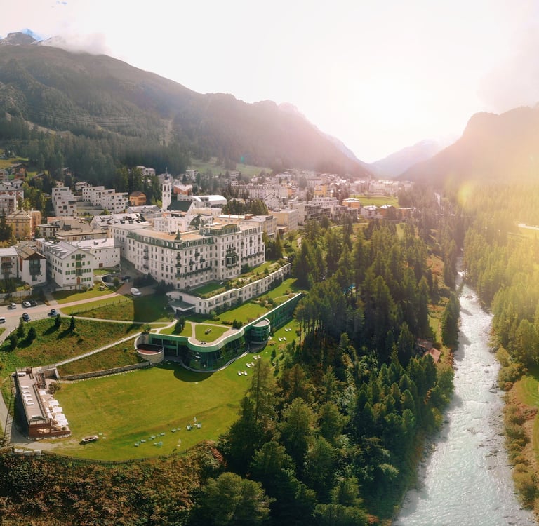 Aerial view of a luxury hotel and alpine village in a sunlit Swiss mountain valley next to a river.