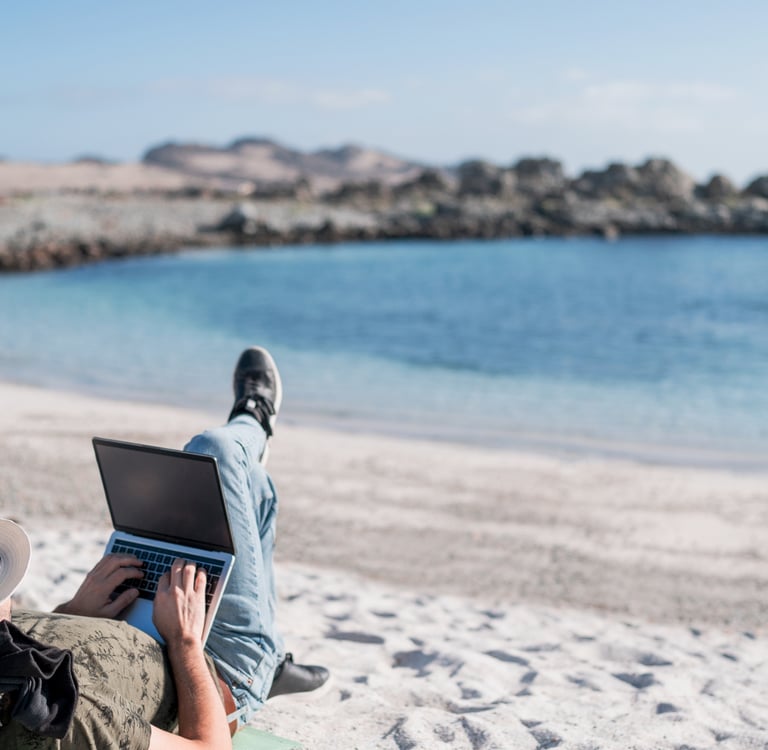 Digital nomad working on a laptop while relaxing on a white sand beach near the blue ocean.