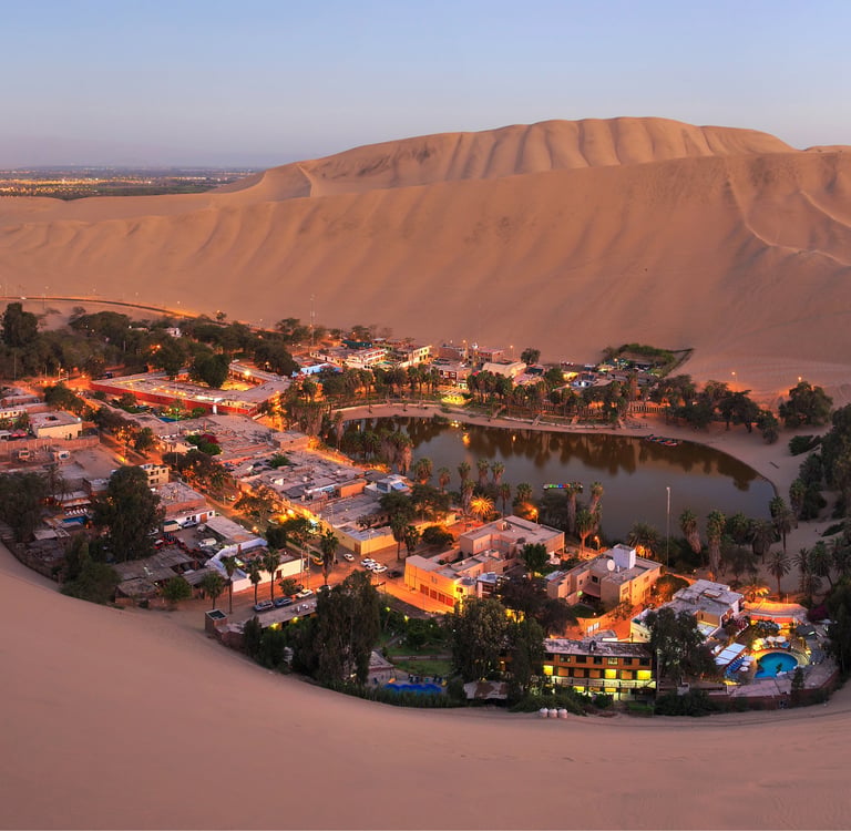 Aerial view of Huacachina desert oasis in Peru with illuminated village buildings and palm trees at dusk.