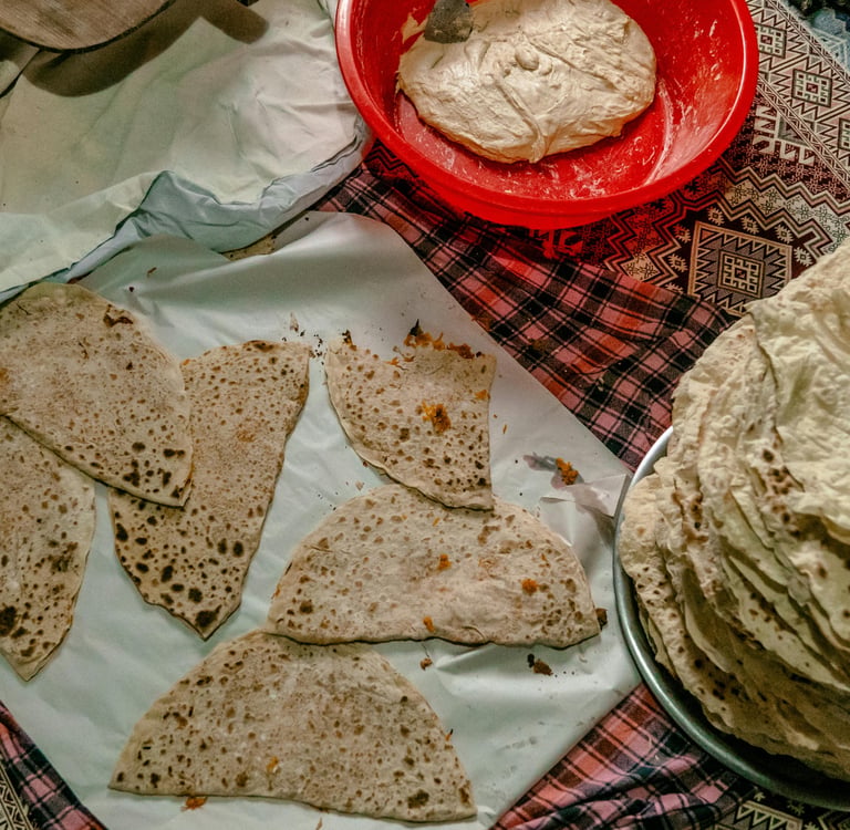 A stack of traditional flatbread and pieces of lavash next to a red bowl of fresh dough on a patterned rug.