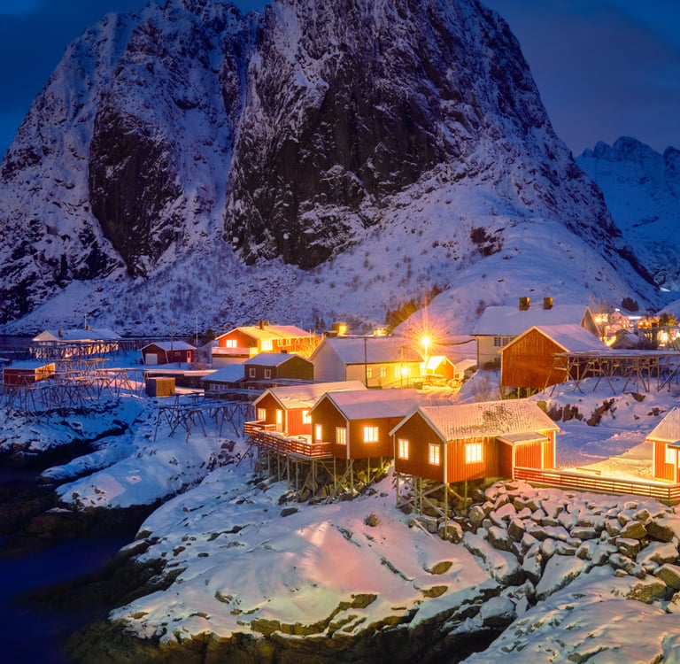 Illuminated red fishing cabins in Reine, Lofoten Islands, nestled under snow-capped mountains at twilight.
