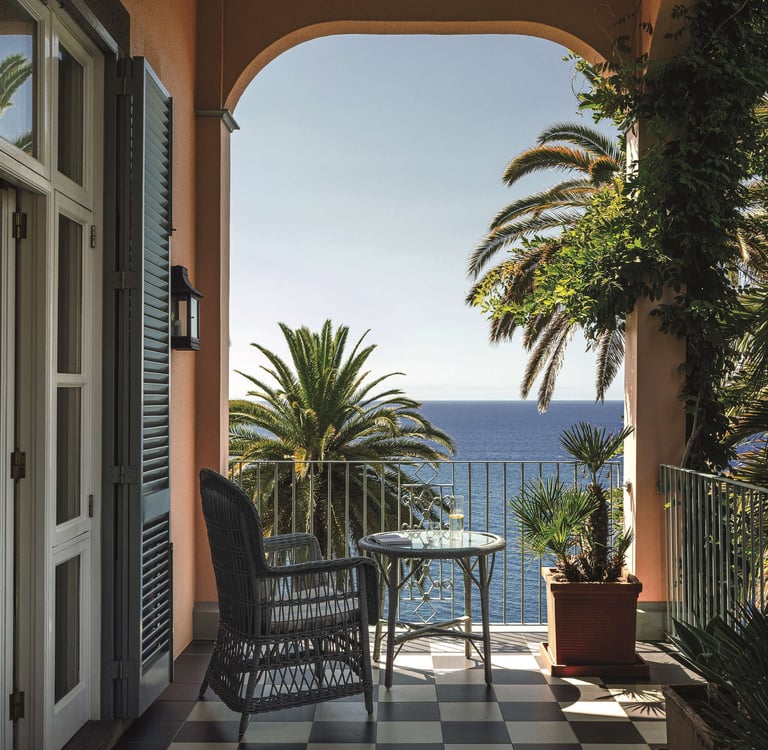 Luxury hotel balcony with ocean views, palm trees, and wicker furniture on a sunny day.