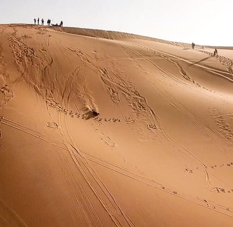 Tourists sandboarding down a high sand dune in the Sahara Desert under a clear sky.