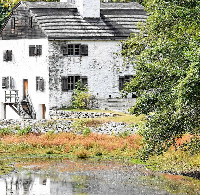 Historic white stone manor house with wooden shutters situated behind a scenic pond and lush green trees.