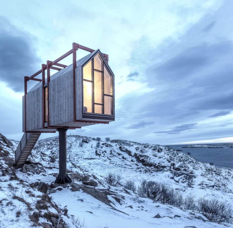 A modern elevated wooden cabin perched on a snowy hillside overlooking a lake under a cloudy sky.