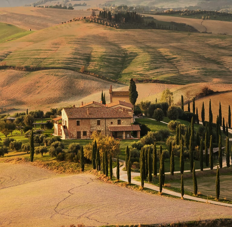 A scenic stone villa on a Tuscan hillside surrounded by cypress trees and rolling golden fields at sunset.