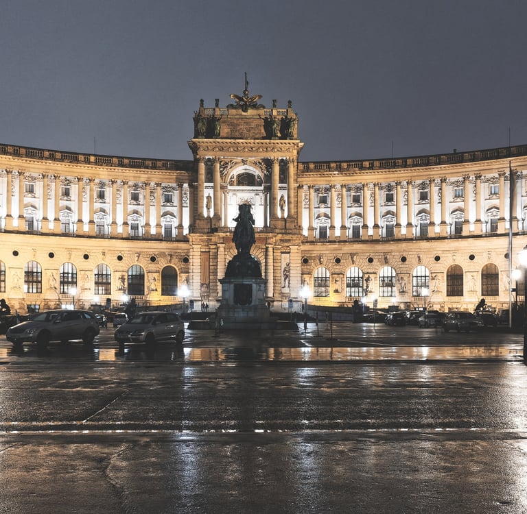 Illuminated facade of Hofburg Palace at night in Vienna, Austria, with wet pavement reflections.
