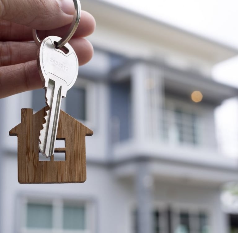 Mão segurando chave com um chaveiro de casa e uma casa atrás