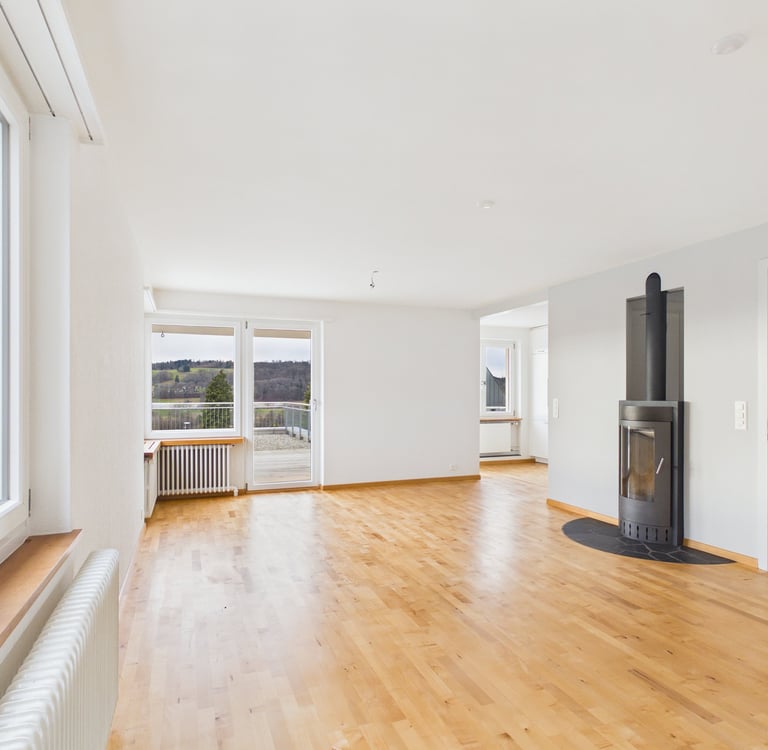 Empty modern living room featuring light hardwood floors, a black wood-burning stove, and large glass balcony doors.