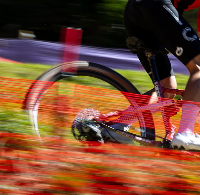 A mountain biker performs a bunny hop over a white bar obstacle on a dirt trail.
