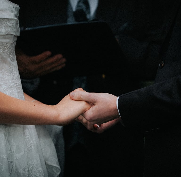 Close-up of a bride in a lace gown and groom in a black suit holding hands during a wedding ceremony.