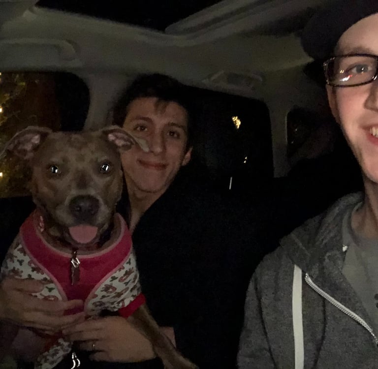 Two people and a dog in a car at night. One person holds a happy dog wearing a sweater. 