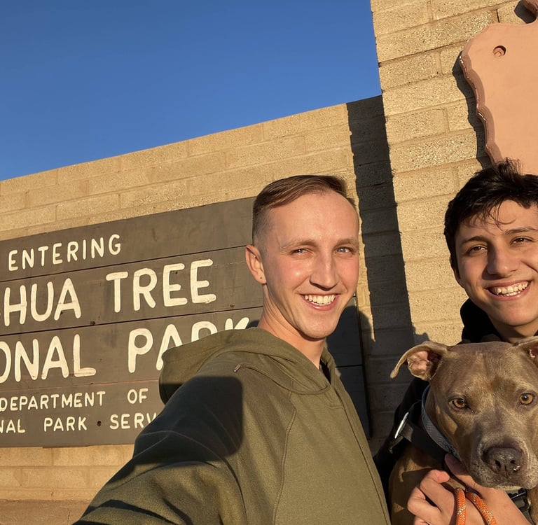 Two people and a dog smile in front of the Joshua Tree National Park entrance sign on a sunny day. 