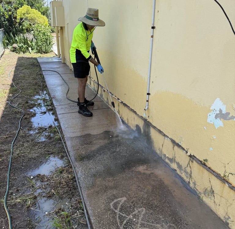 A professional pressure washing technician cleaning a concrete sidewalk and exterior wall.