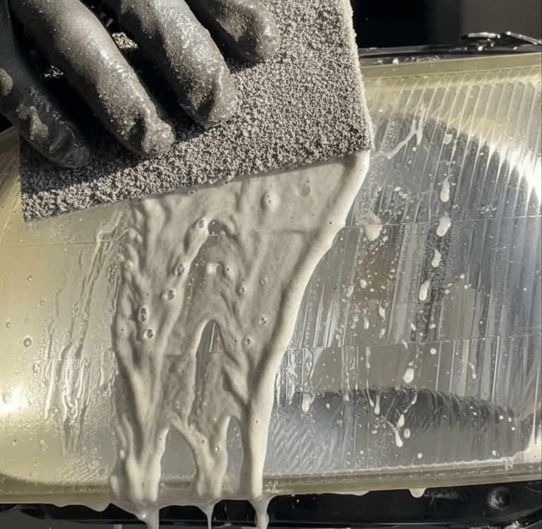 A gloved hand uses a sanding sponge to restore a yellowed car headlight with a cleaning solution.