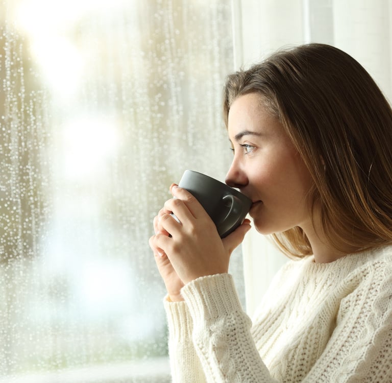 a woman drinking from a mug sitting by a window and its raining outisde