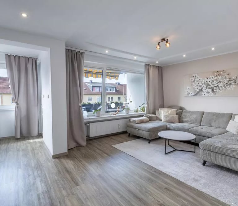 Modernes Wohnzimmer mit grauem Ecksofa, Holzboden und großen Fenstern mit neutralen Vorhängen.