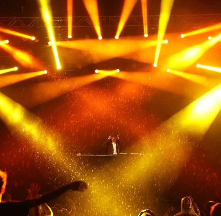 A DJ performs at a live music concert with dramatic golden stage lighting and special effects.