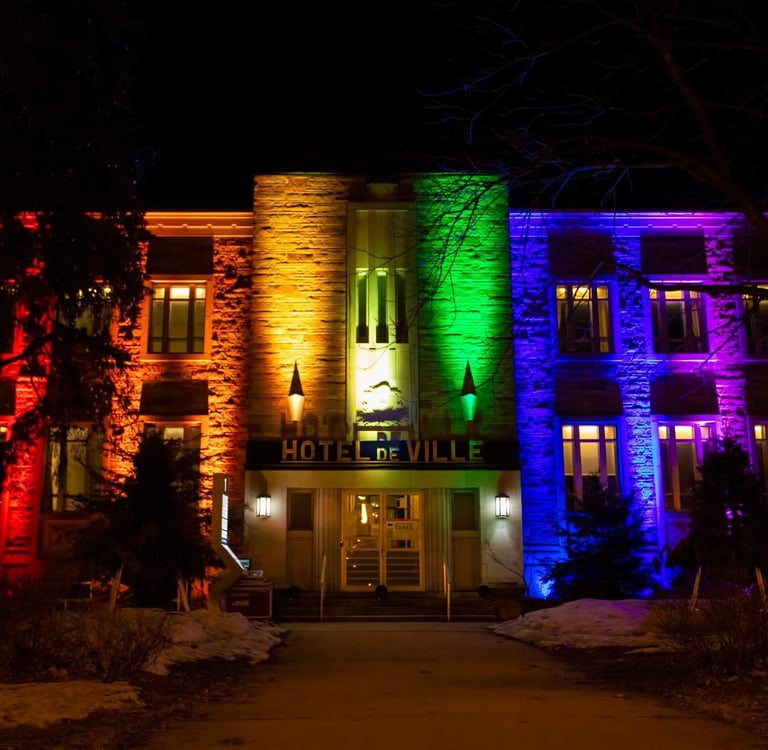 The Hotel de Ville building illuminated with vibrant rainbow pride colors at night.