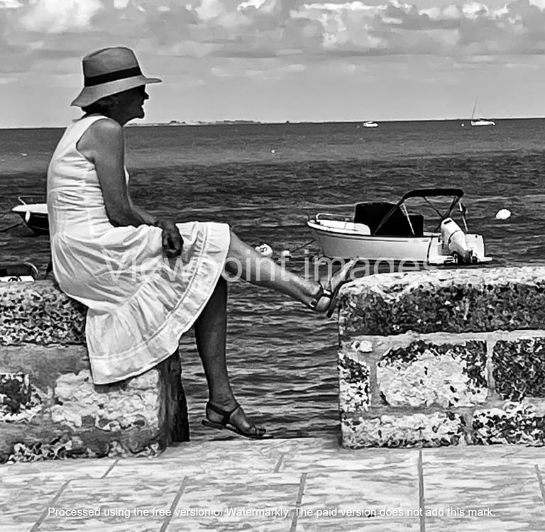 Black and white photo of a woman in a sun hat and white dress sitting on a stone wall by the sea.