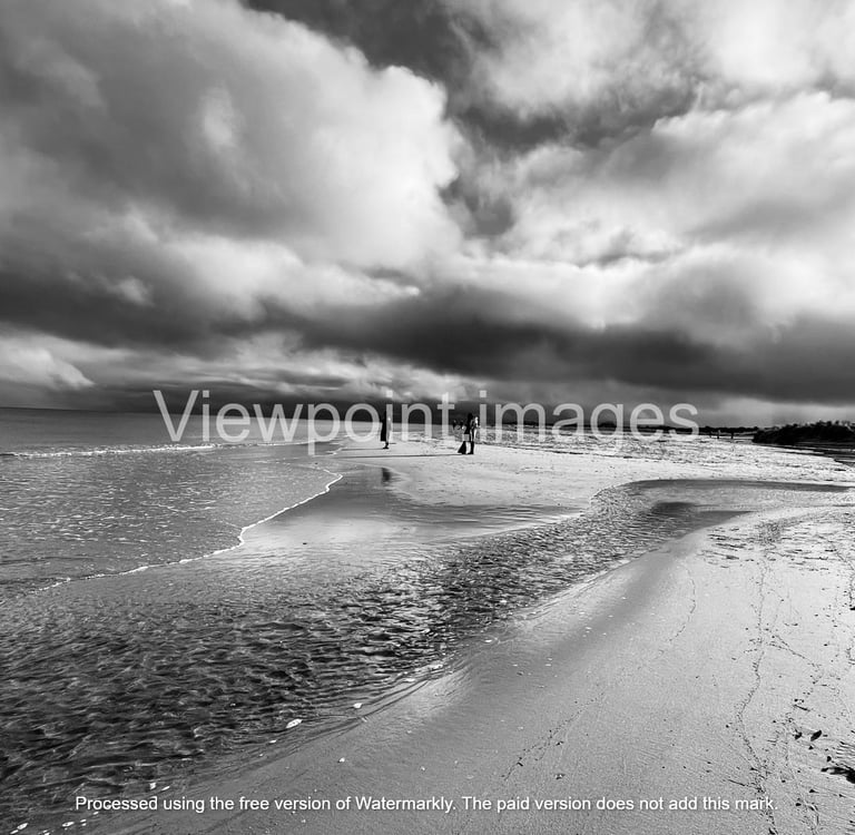 Dramatic black and white landscape of a sandy beach with dark storm clouds reflecting on the ocean tide.
