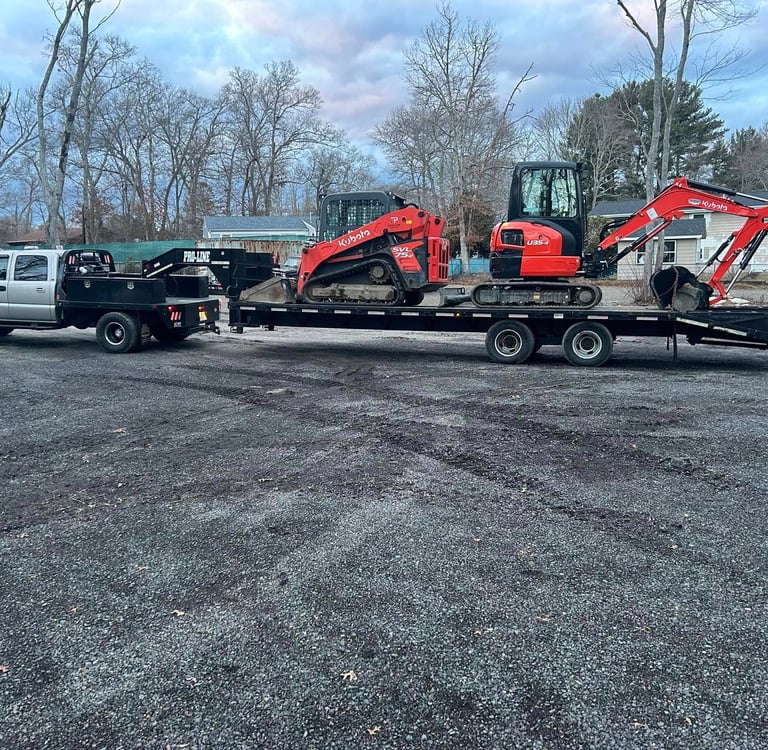A silver flatbed truck towing a Kubota skid steer and mini excavator on a gooseneck trailer.