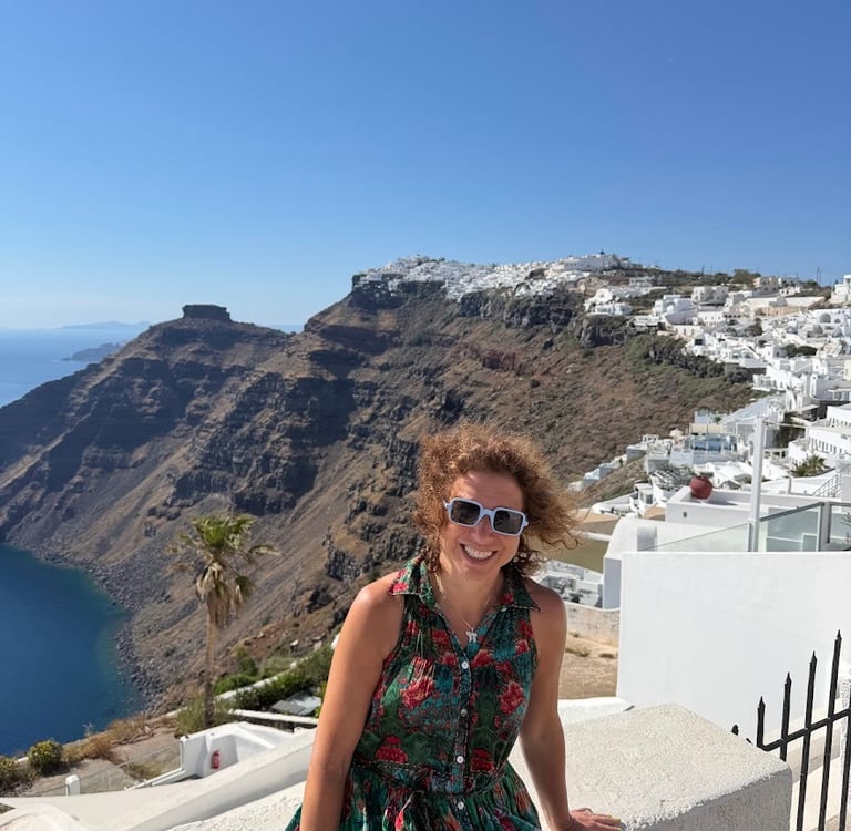 A smiling woman in a floral dress overlooking the Santorini caldera and white buildings of Imerovigli, Greece.