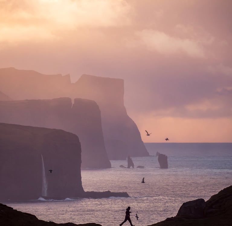 Man running along Kallur lighthouse cliffs with dramatic drops and ocean below, wild Faroe Islands landscape