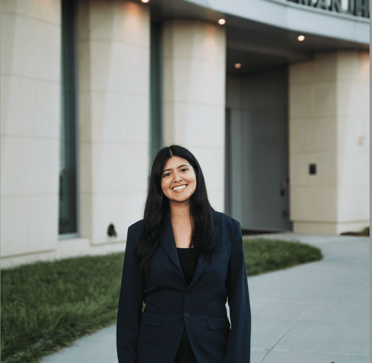 Professional woman in a navy blazer smiling in front of the Hartman Hall building on campus.