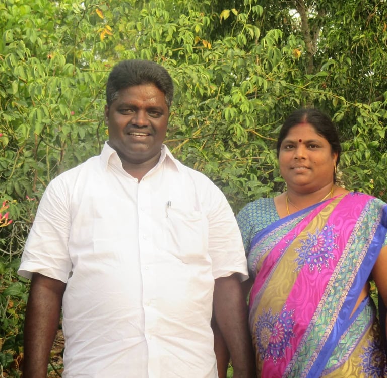 A smiling Indian couple posing together outdoors against a lush green leafy background.