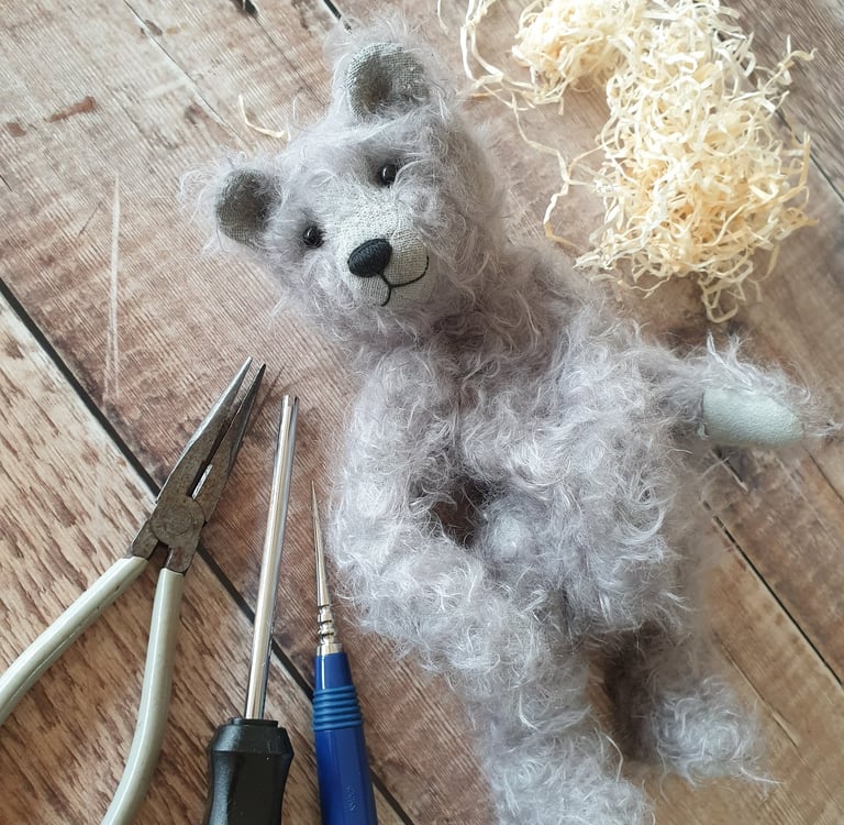 a partly made handmade teddy bear in grey mohair with a wistful face and surrounded by tools on a table