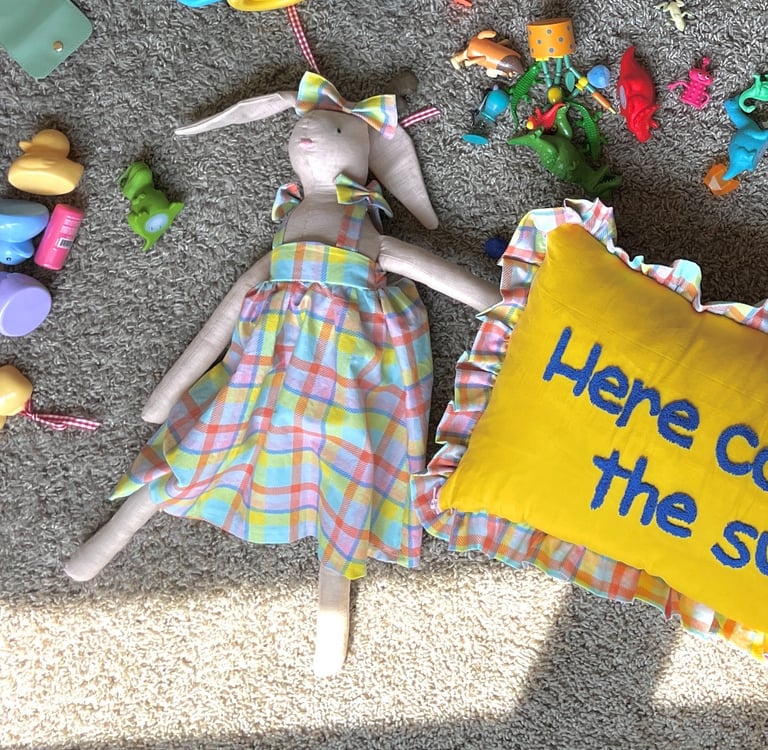 Handmade cloth bunny doll in a plaid dress lying on a carpet with kids toys and a yellow pillow.