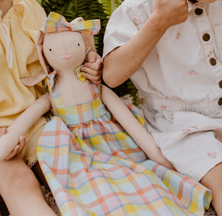 Two young children sitting together holding a handmade plush bunny doll wearing a colorful plaid dress.