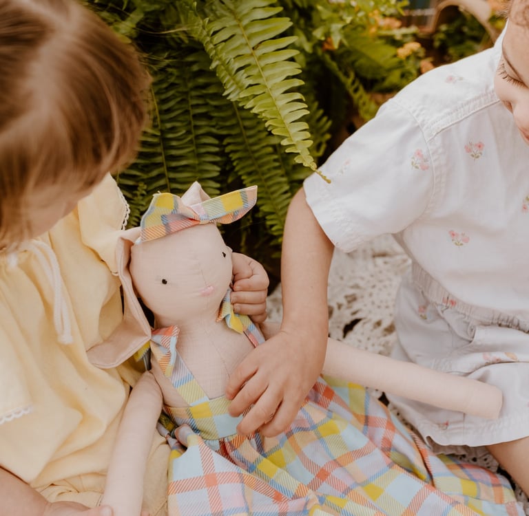 Two young children playing with a soft linen bunny doll outdoors near a lush green fern.