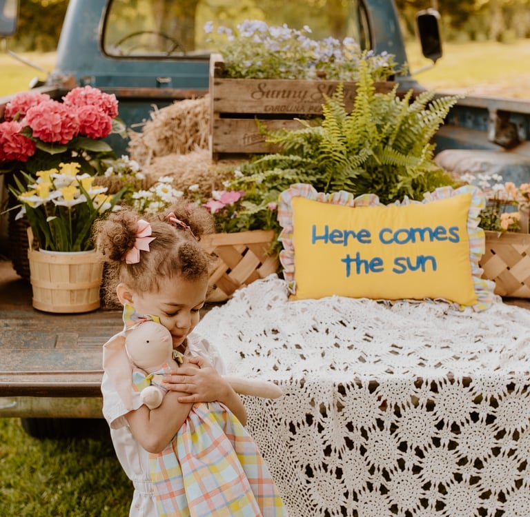 A toddler holding a doll stands near a vintage blue truck bed filled with spring flowers and a yellow sun pillow.