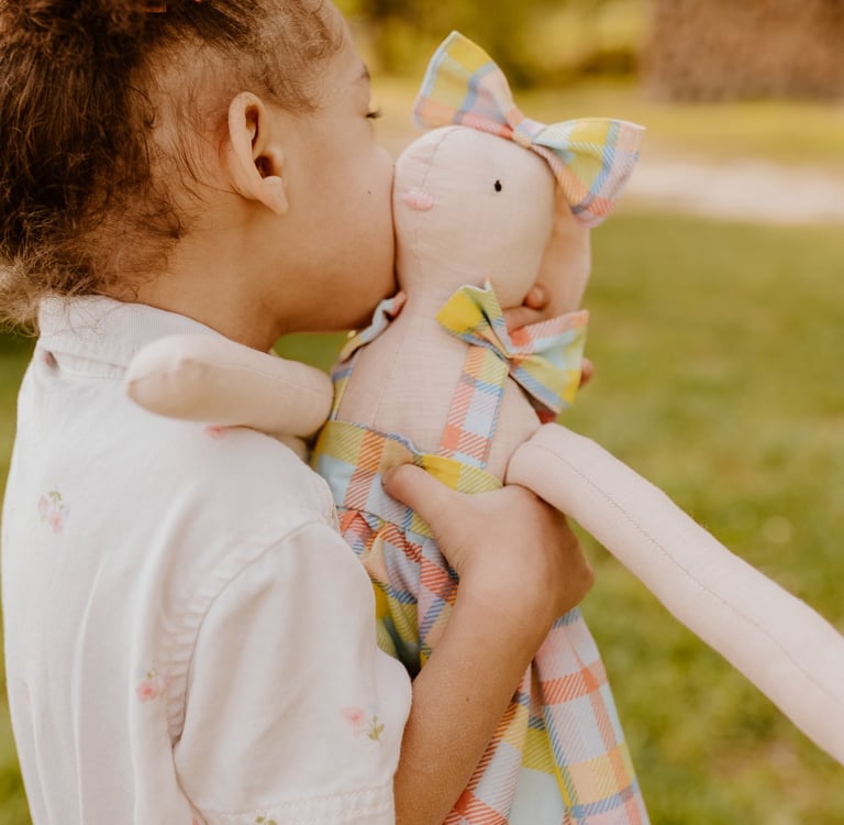 A young girl with curly hair hugs and kisses a handmade linen bunny doll outdoors.