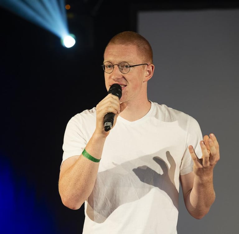 Przemysław Żuk, kurz Przemek, steht auf einer Bühne und spricht in ein Mikrofon. Er gestikuliert. Im Hintergrund Bühnenlicht.