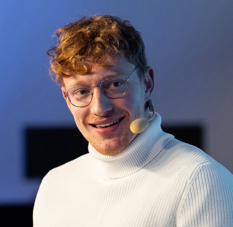 Moderator Przemysław Żuk, kurz Przemek Żuk, moderiert lächelnd eine Paneldiskussion. Er trägt ein Headset-Mikrofon.