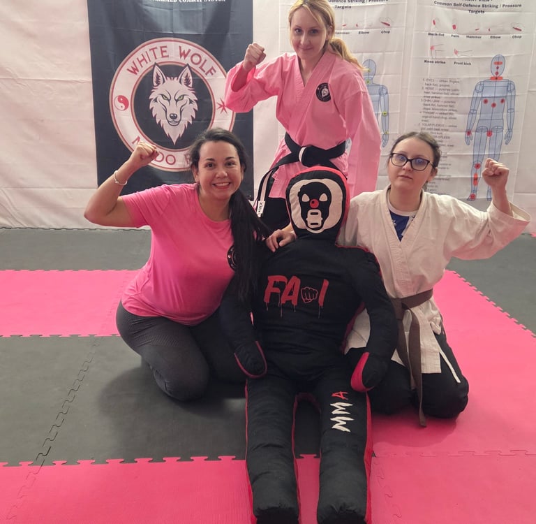 Three female martial artists posing with an MMA training dummy on a pink gym mat.