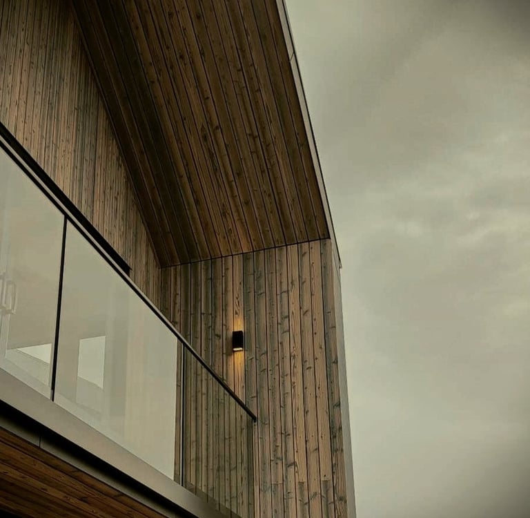 Modern balcony featuring vertical cedar wood siding, glass railing, and exterior wall lighting under a moody sky.