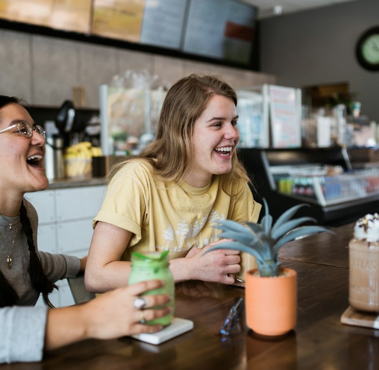 Dos jóvenes sonríen y conversan en una cafetería moderna, disfrutando bebidas y representando la cul