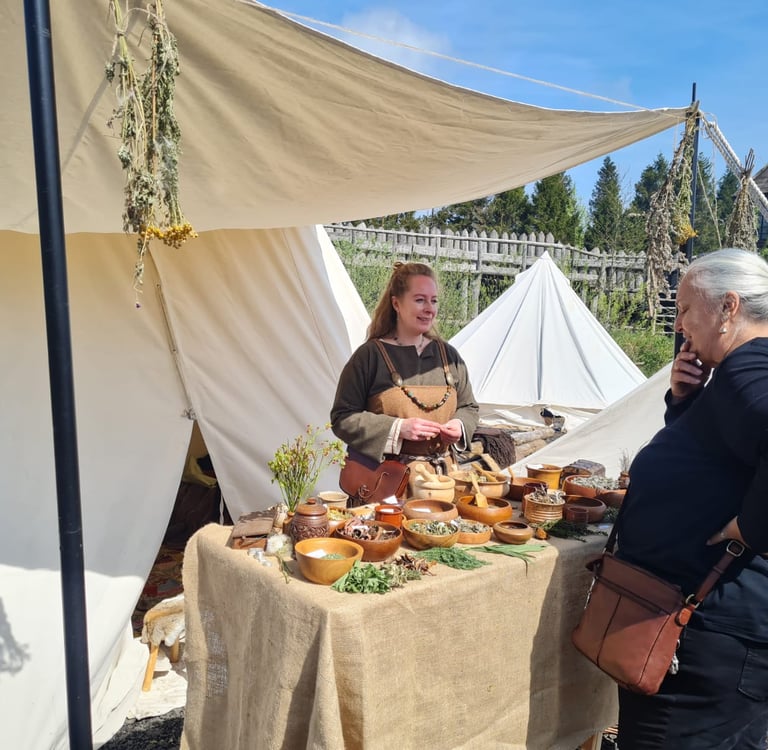 a woman dressed in viking clothing standing behind a table of herbal remedies talking  with visitors