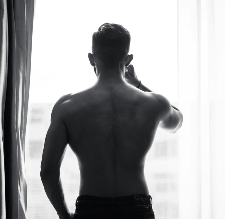 Silhouette of a muscular shirtless man looking out a bright window in black and white.