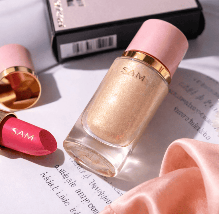 Iluminador líquido dorado de SAM Cosméticos junto a labial sobre fondo elegante.